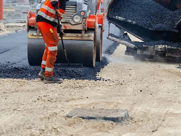 Worker in hi-vis clothing raking fresh asphalt in front of a roller and a paver